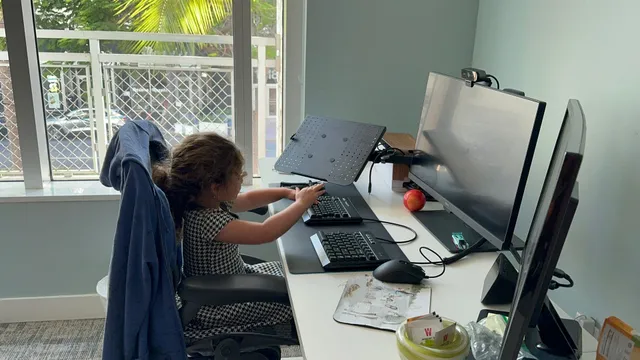 One of AJ's daughters sitting at his office desk, reaching for the keyboard between two monitors.