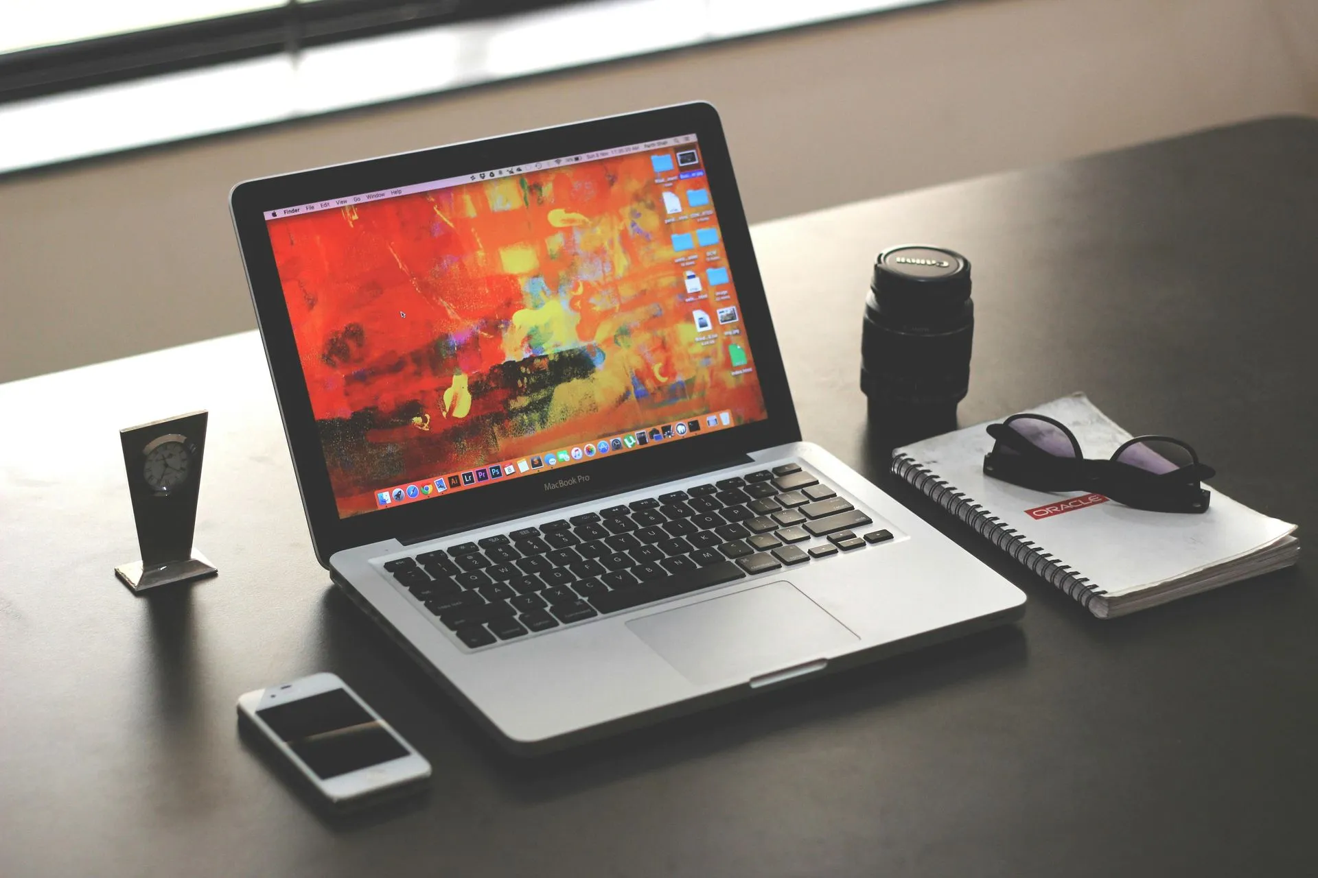 A laptop, smartphone, lens, and notebook with sunglasses resting on a dark desk near a window.