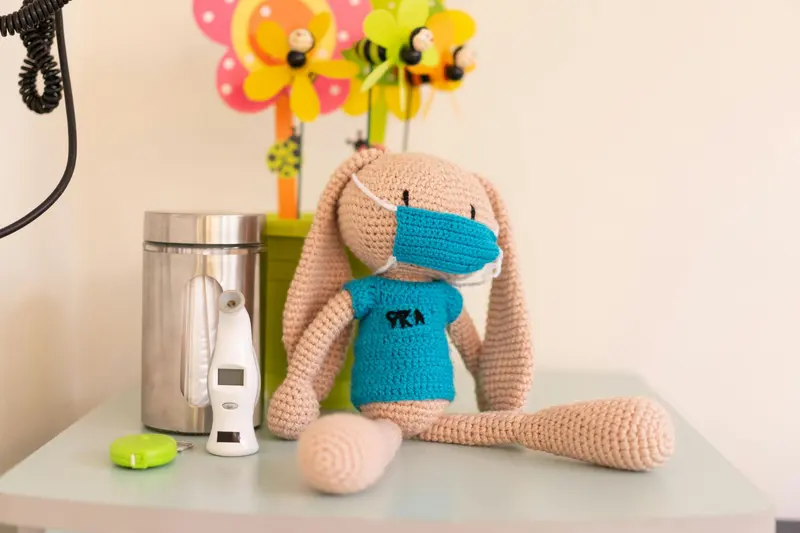 A crocheted toy rabbit in a doctor's scrubs and mask sits on a child's nightstand beside a digital thermometer.