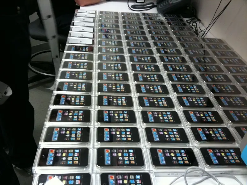 Dozens of identical smartphones laid out in neat rows on a table in an indoor setting.