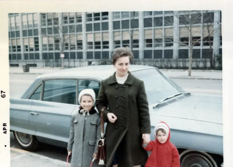 A 1967 photograph of a young woman in a dark coat holding two small children's hands beside a parked car.
