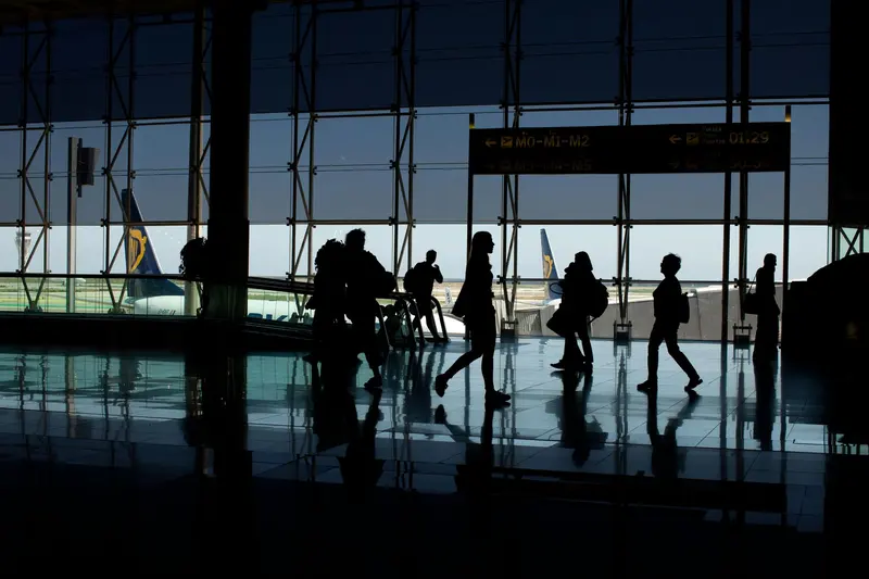 Travellers silhouetted against the windows of an airport terminal, with planes parked outside.