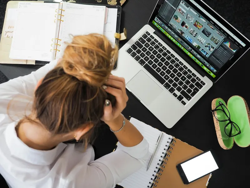 Someone at a cluttered desk with their head in their hands, a laptop and open planner in front of them.
