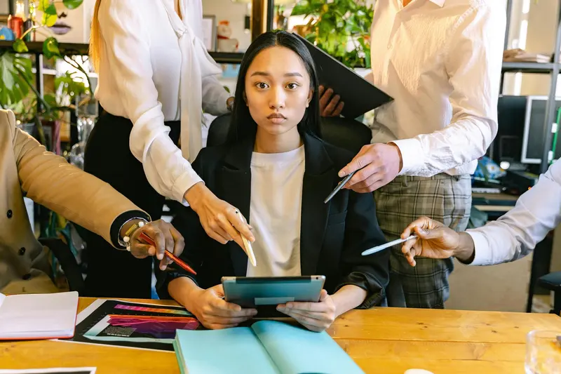 A woman at a desk holding a tablet while colleagues crowd in, thrusting pens and papers at her.