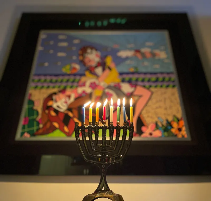 A brass menorah with eight coloured candles burning, lit in front of a framed piece of colourful folk art.
