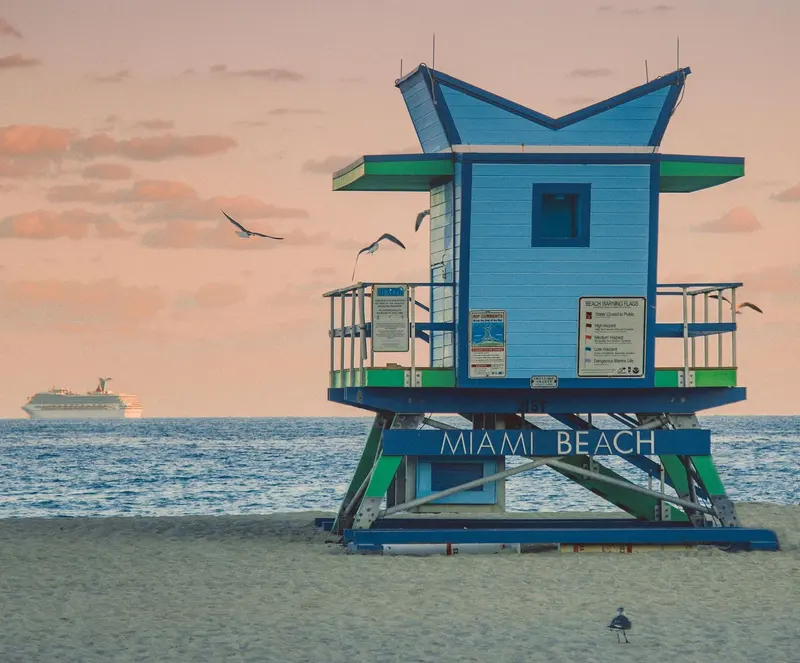 A blue Miami Beach lifeguard tower at dusk, gulls overhead and a cruise ship on the horizon.