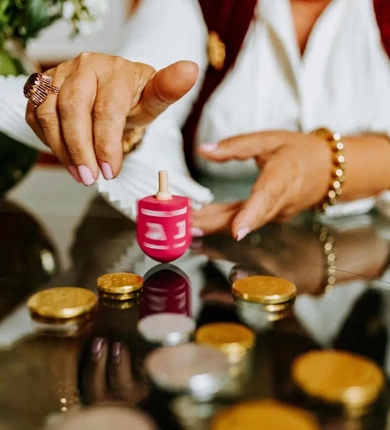 Hands spinning a pink dreidel on a glass table scattered with gold chocolate coins.