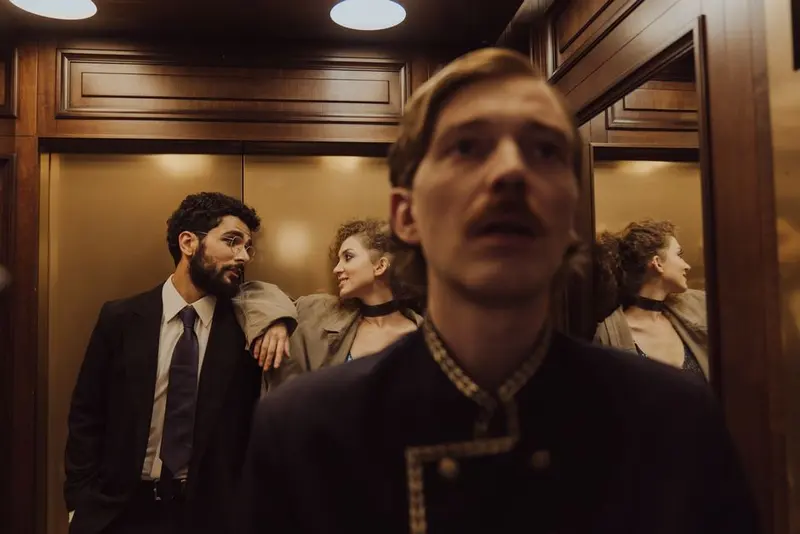 Three people standing in a wood-panelled elevator, a hotel bellhop in uniform blurred in the foreground.