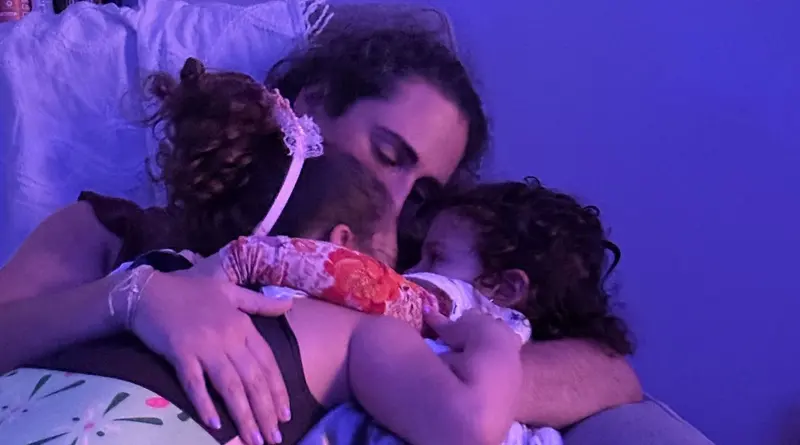 A parent lying in dim purple light with two small daughters curled up asleep against them.