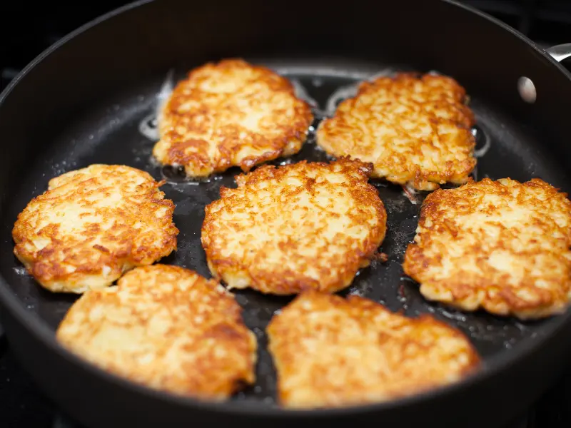 Seven latkes frying in oil in a black skillet, browned and crisp at the edges.