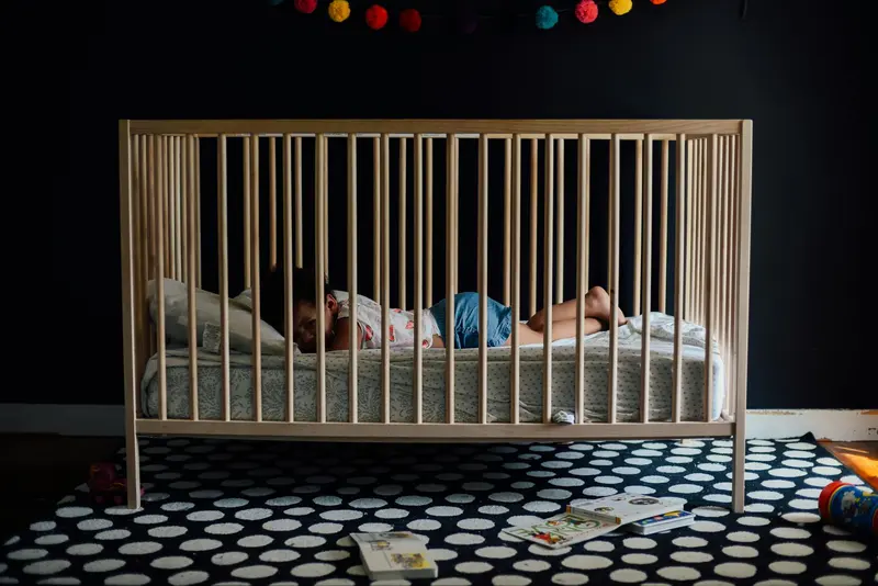 A toddler lying awake in a wooden crib in a darkened room, books scattered on the rug below.