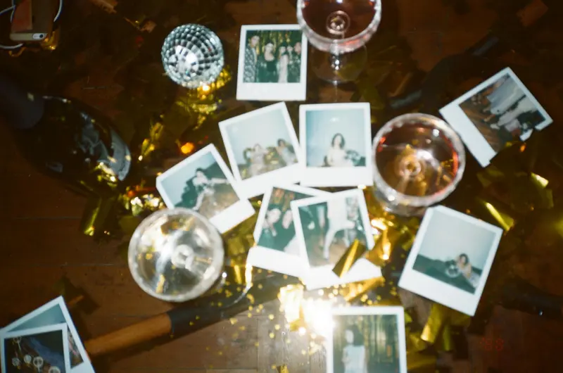Polaroid photos scattered across a wooden floor among gold confetti, wine glasses, and a small disco ball.