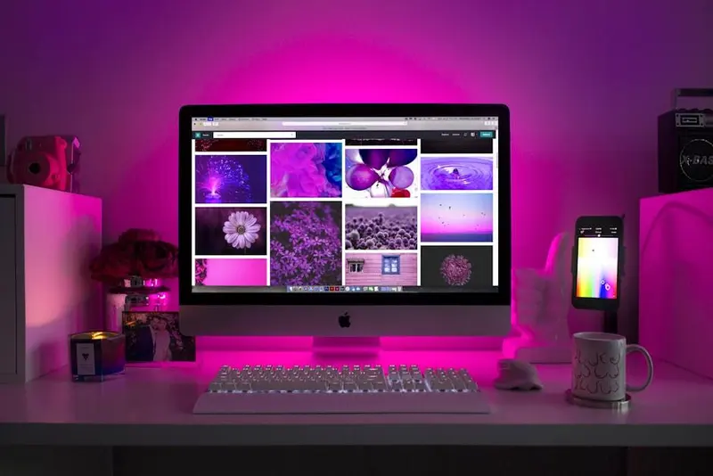 A desktop computer on a white desk showing a purple photo grid, the whole room lit by pink neon.