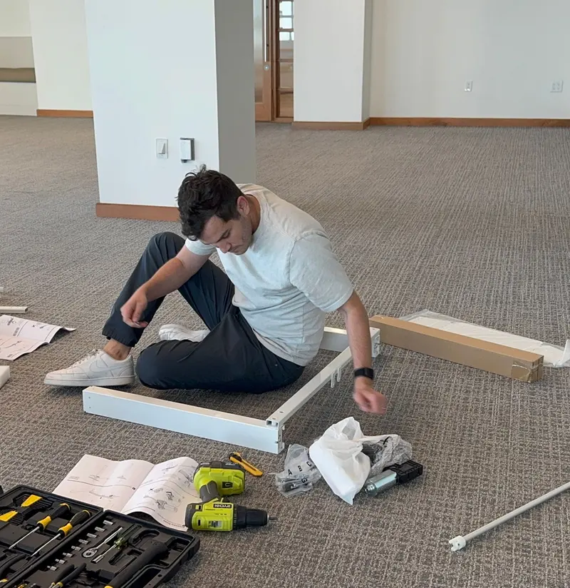 A man sitting on an office floor assembling flat-pack furniture, tools and instructions spread around him.