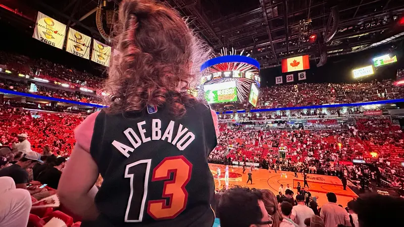 A child in an Adebayo number 13 Heat jersey watching a game from the stands at a packed arena.
