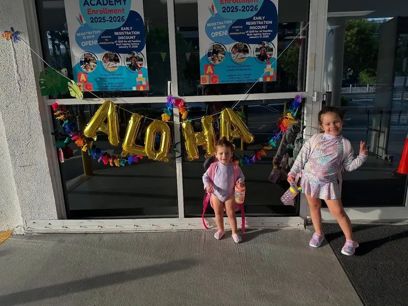 Yael and Diana posing outside their school under a gold ALOHA balloon banner, backpacks on.