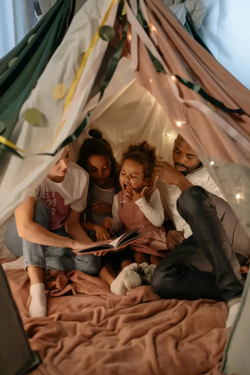 A family sitting together inside a blanket fort strung with fairy lights, reading a picture book.