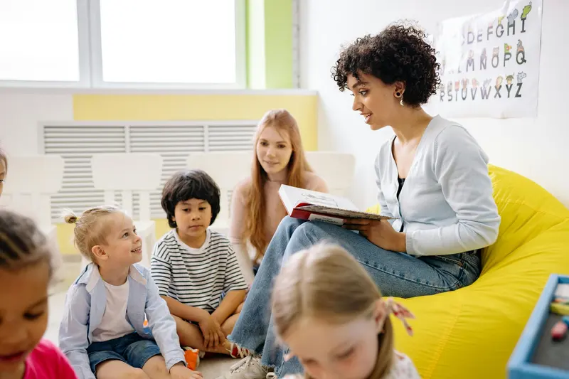 A teacher sitting on a yellow beanbag reading a picture book to a circle of young children.