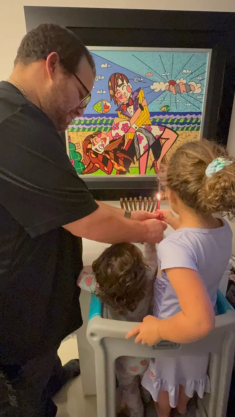AJ helping his daughter light the menorah while her younger sister watches from a high chair.