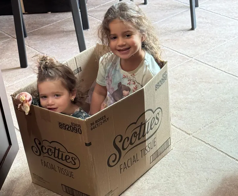 Yael and Diana sitting inside a large cardboard box on the kitchen floor, grinning at the camera.