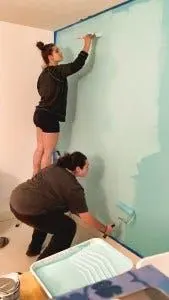 Two people painting an interior wall pale teal, one on a step stool and one working with a roller tray.