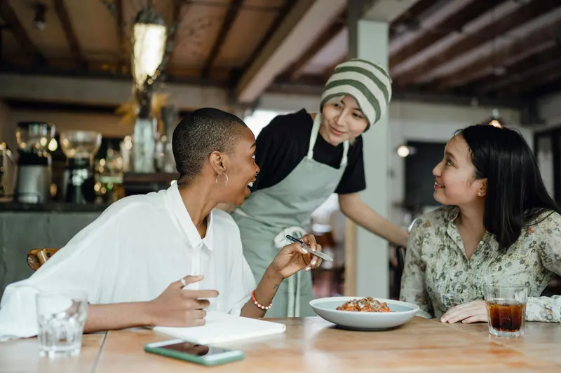 A server in an apron leaning in to talk with two smiling diners at a restaurant table.
