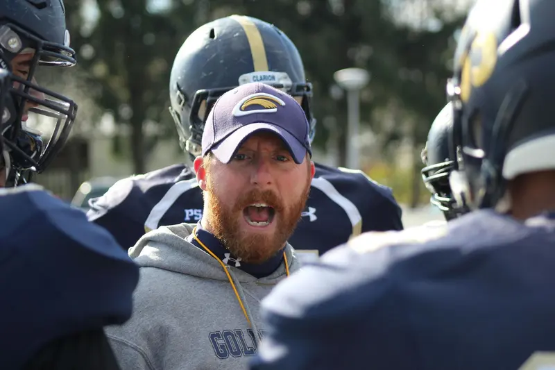 A football coach in a cap and hoodie shouting instructions inside a huddle of helmeted players.