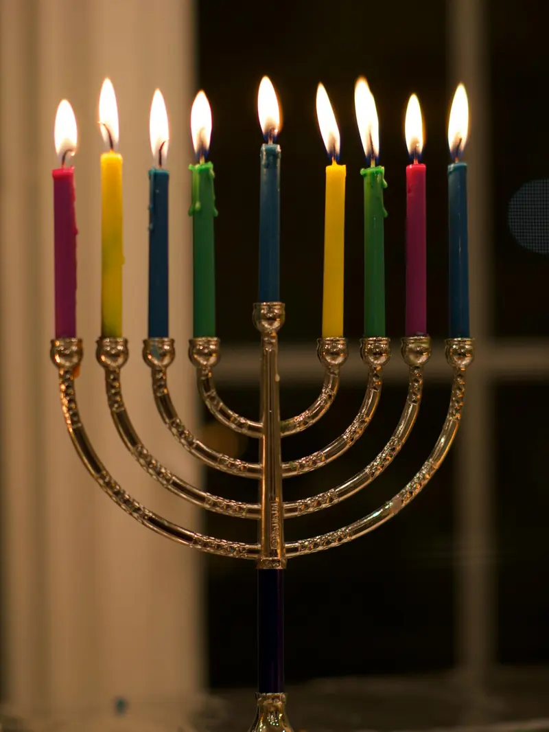 A silver menorah with nine brightly coloured candles lit, glowing against a dark window.