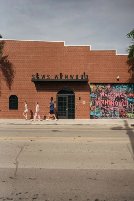 Pedestrians walking past a Wynwood warehouse painted with a mural reading 'Welcome to Wynwood'.
