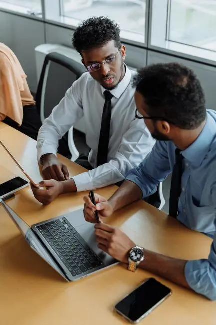 Two colleagues in shirts and ties leaning over a laptop in a meeting room, mid-discussion.