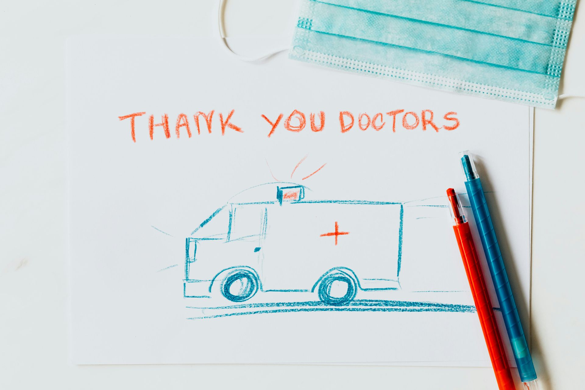 A child’s crayon drawing of an ambulance captioned ‘Thank you doctors’, beside a surgical mask and pens.
