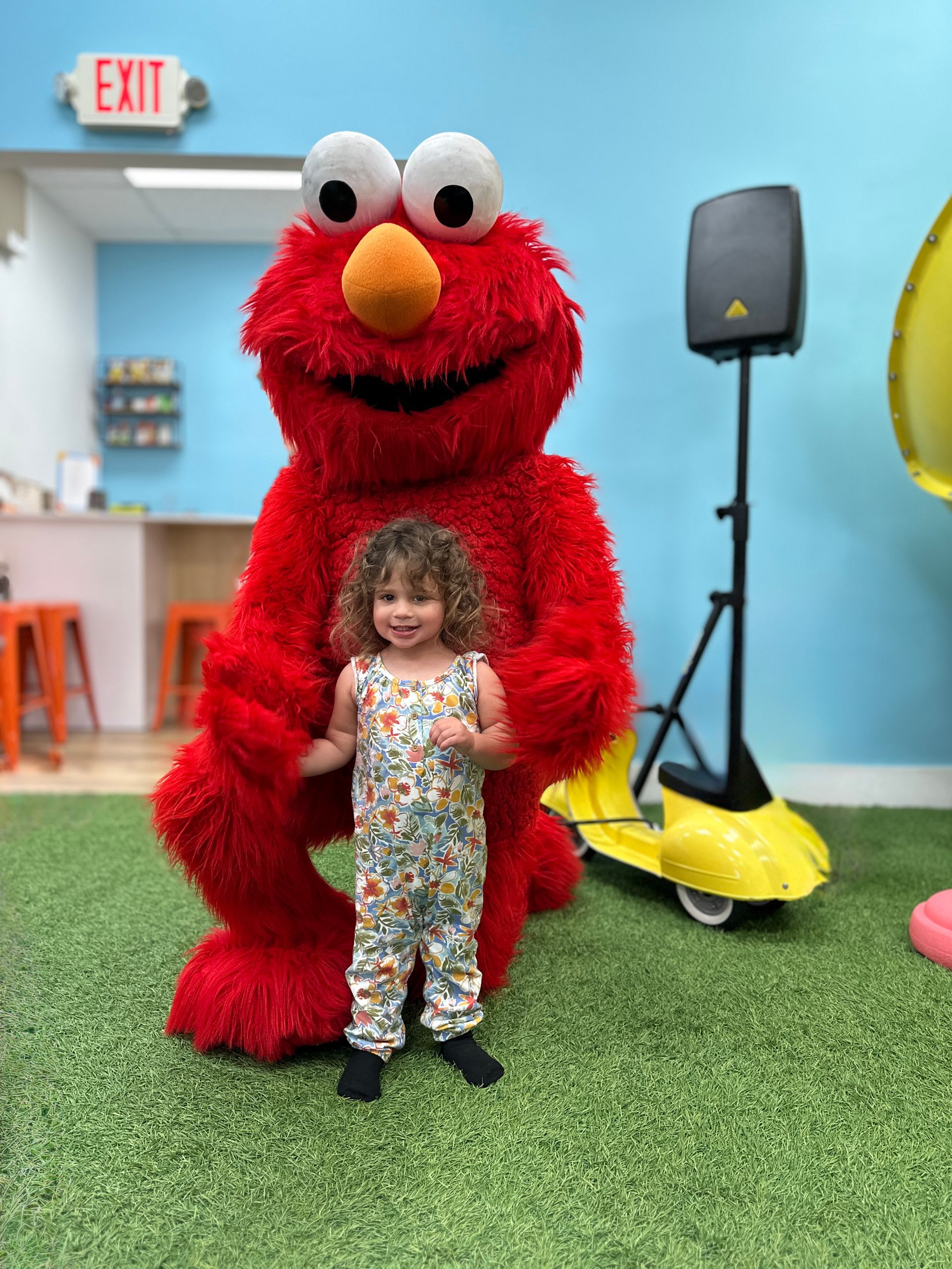 One of AJ’s daughters standing in front of a life-size Elmo character at an indoor play space.
