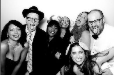 A black and white photo booth strip: six family members crowded together pulling faces in novelty horns and bunny ears.